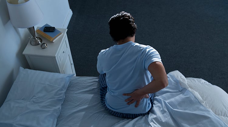 A person sitting on a bed holding their back in pain.